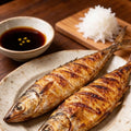 Two charcoal-grilled mackerel with crisp scored skin on a plate with a dipping sauce